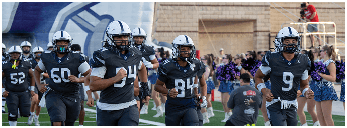 People in football uniforms are jogging on a field with cheerleaders and a photographer in the background. Descripci n generada con IA
