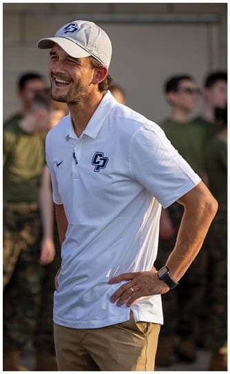 A person wearing a white polo shirt and matching cap with a logo is smiling and standing with hands on hips while others in military-style clothing stand blurred in the background. Descripci n generada con IA