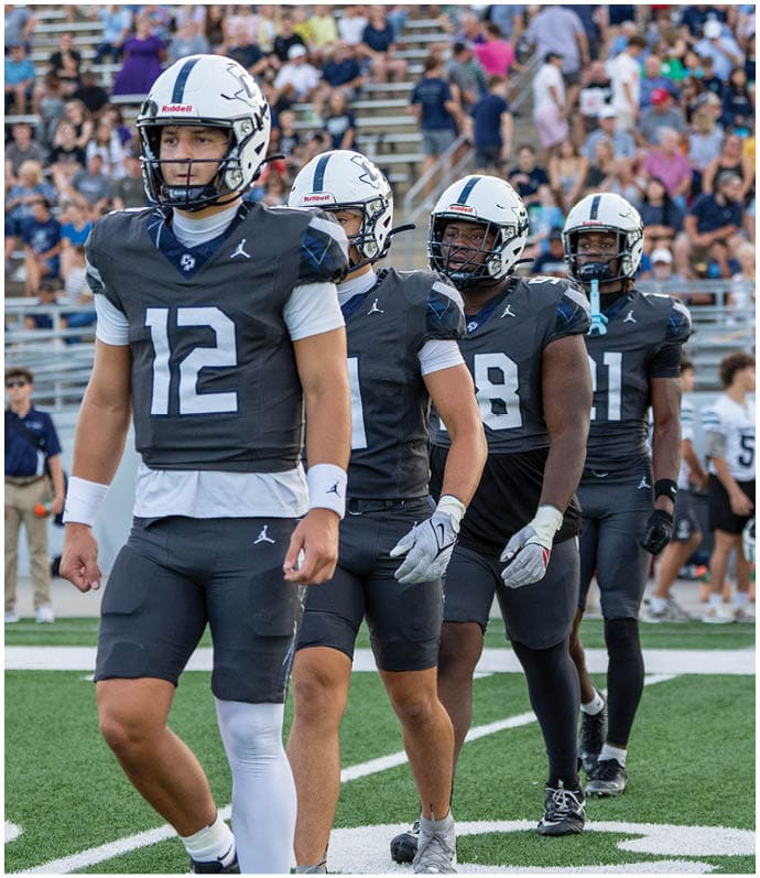 Two people wearing dark football uniforms and white helmets stand on a field with blurred spectators in the background. Descripci n generada con IA