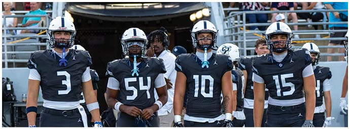 Four people wearing black football uniforms with helmets standing on a field with bleachers and spectators in the background. Descripci n generada con IA
