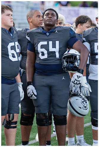 A person wearing a dark gray football uniform with the number 64 is standing while holding a white football helmet and wearing white gloves. Descripci n generada con IA