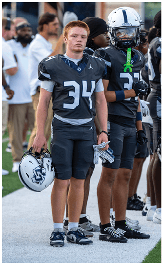 A person with short red hair wearing a dark football uniform with the number 34 stands holding a helmet and gloves while other players and a blurred person are in the background. Descripci n generada con IA