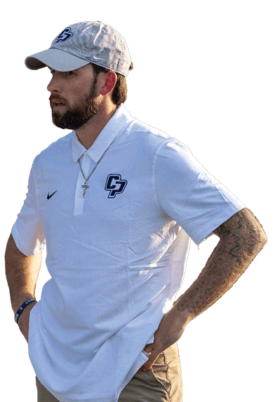 A person wearing a white polo shirt with a logo, beige pants, a light-colored cap, and a necklace is standing with hands on hips outdoors. Descripci n generada con IA