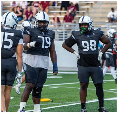 Two people wearing white football helmets are in the foreground with blurred spectators sitting on bleachers in the background. Descripci n generada con IA