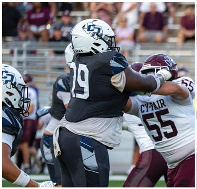 Two people in football uniforms are engaged in a play on the field, with one wearing a black jersey numbered 79 and the other in a white jersey numbered 55. Descripci n generada con IA