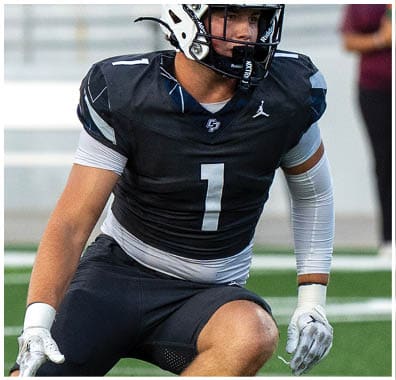 A person wearing a black football uniform with the number one and a white helmet is on a football field with empty bleachers in the background. Descripci n generada con IA