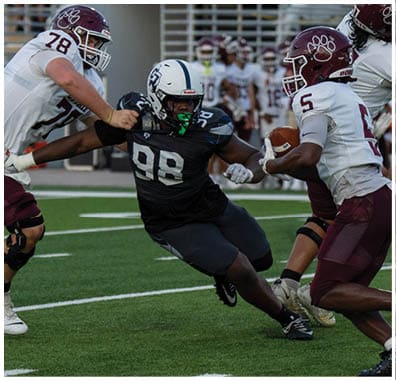A person wearing a black football uniform with the number 98 is being tackled by two people in white and maroon football uniforms during a game on a green field. Descripci n generada con IA