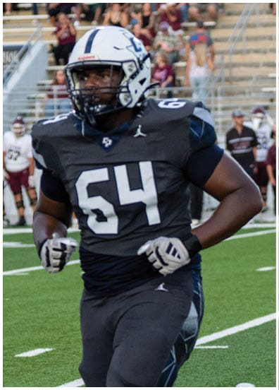 A person wearing a dark football jersey with the number 64 and white gloves is running on a field. Descripci n generada con IA