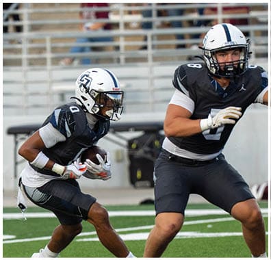 Two people in football uniforms are on a field with one holding a football and the other preparing to block or assist. Descripci n generada con IA