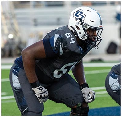 A person in a football uniform with a helmet and knee braces is crouched in a ready stance on a green field with white yard lines. Descripci n generada con IA