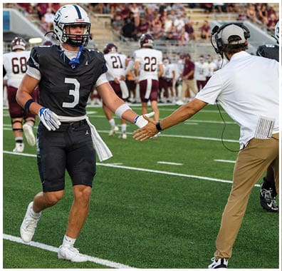 A person wearing a black football uniform with the number three is giving a high five to a person in a white shirt and khaki pants on a football field during a game. Descripci n generada con IA