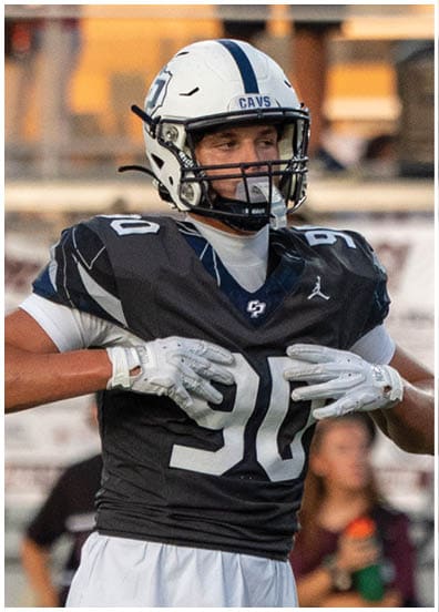A person wearing a football uniform and helmet is adjusting their gloves on a sports field with blurred spectators in the background. Descripci n generada con IA
