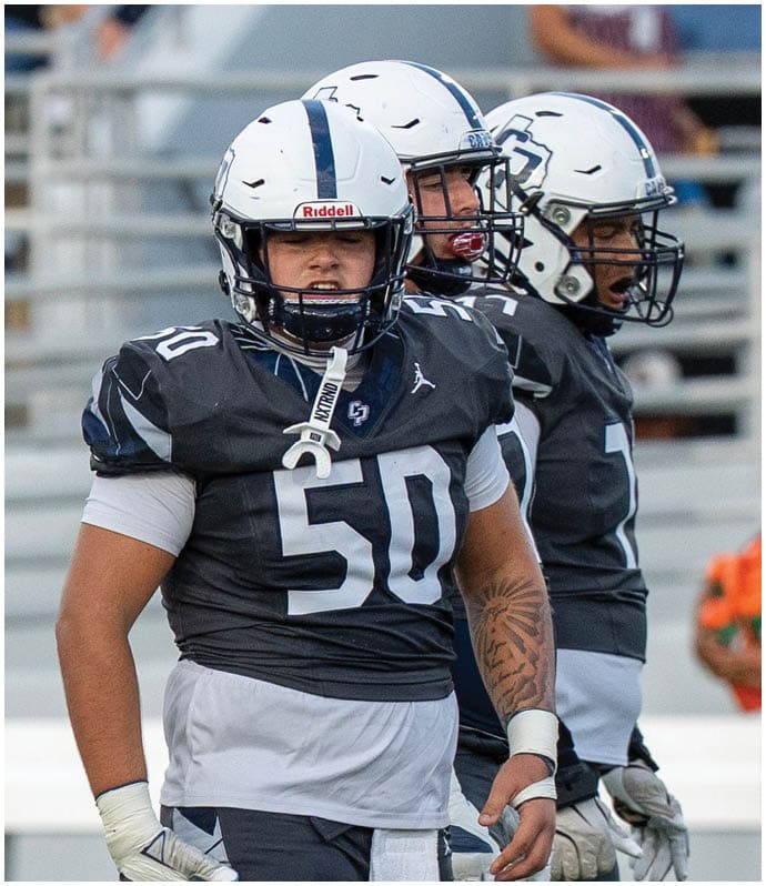Three people wearing football helmets and black and white uniforms stand on a field with one person in the foreground wearing number 50. Descripci n generada con IA