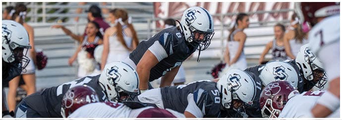 A person in a football uniform and helmet is on the field with blurred people and a banner reading “CFHS Brigade" in the background. Descripci n generada con IA