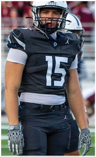 A person wearing a black football uniform with the number 15 and a white helmet stands on a football field with blurred spectators in the background. Descripci n generada con IA