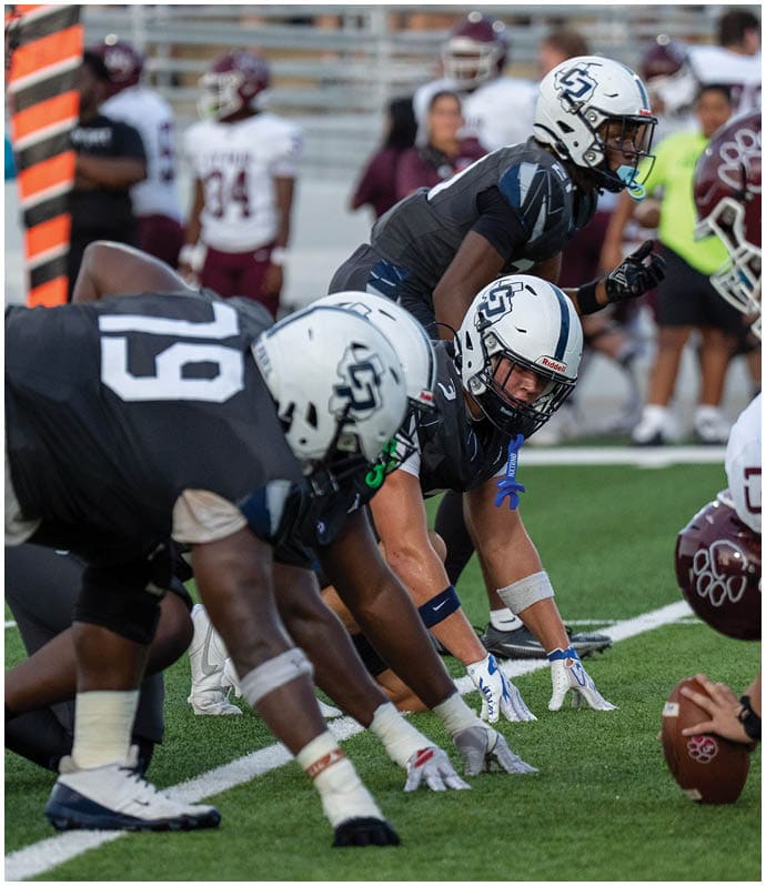 Two opposing American football teams are lined up at the line of scrimmage on a green field, preparing for the next play. Descripci n generada con IA