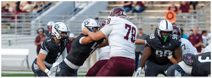 People wearing football uniforms are engaged in a play on a football field with one person in a white jersey blocking others in dark jerseys. Descripci n generada con IA