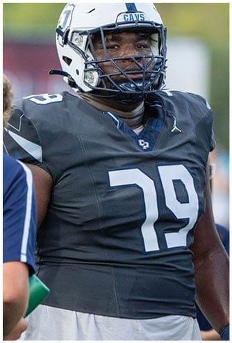 A person wearing a dark football jersey and a white helmet with a faceguard is on a football field. Descripci n generada con IA