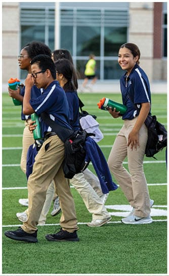 People wearing matching navy blue shirts and khaki pants are walking outdoors on a sports field, some holding green water bottles and one person smiling. Descripci n generada con IA