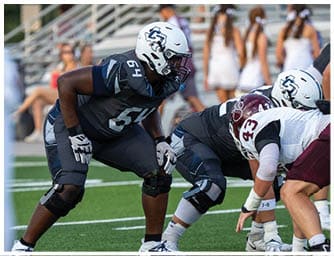 Two people in football uniforms and helmets are crouched in a ready position on a grassy field during a game. Descripci n generada con IA