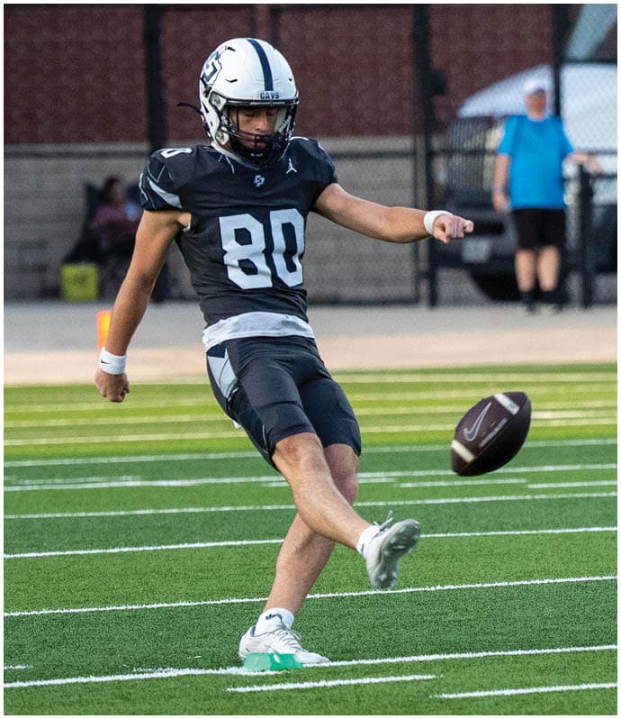 A person wearing a black football uniform with the number 80 is kicking a football on a green field with white yard lines. Descripci n generada con IA