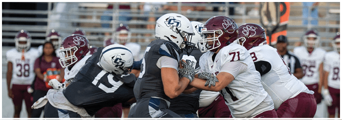 People wearing helmets and football uniforms are engaged in a close physical play on a football field with blurred spectators in the background. Descripci n generada con IA