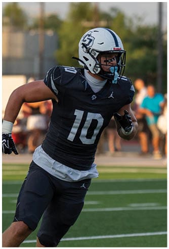 A person wearing a dark sports uniform and a white helmet is preparing to throw a football on a field. Descripci n generada con IA