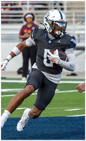 A person in a dark football uniform is running with the ball while another person in a white and maroon football uniform attempts to tackle them on a football field. Descripci n generada con IA