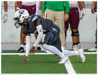 A person in a dark sports uniform and helmet is crouched low on a football field near the sideline with several people standing in the background. Descripci n generada con IA