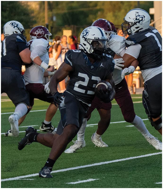 A person wearing a dark football uniform with the number 23 is running with the ball on a football field while other players in dark and white uniforms are nearby. Descripci n generada con IA