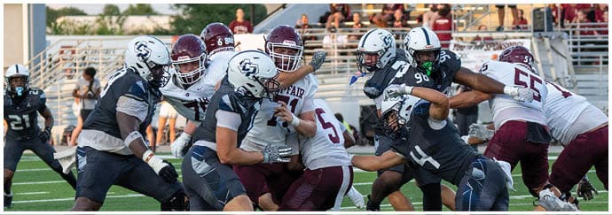 People in football uniforms are engaged in a physical play on a field with spectators in the background. Descripci n generada con IA
