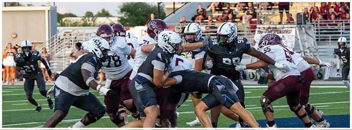 People wearing helmets and football uniforms are engaged in a play on a green football field with bleachers and spectators in the background. Descripci n generada con IA