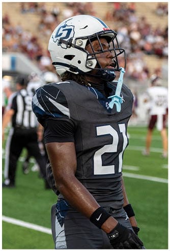 A person wearing a dark football uniform with the number 21 and a white helmet stands on a football field with blurred players and spectators in the background. Descripci n generada con IA