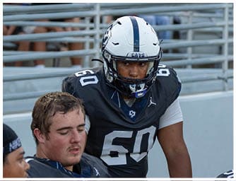 A person wearing a football helmet and jersey number 60 is standing next to another person in a football uniform sitting on a bench in a stadium. Descripci n generada con IA