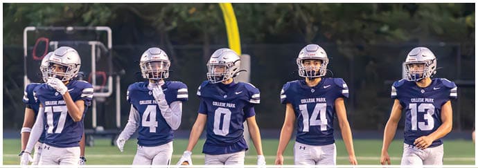Four people wearing football helmets and white Bishop Verot jerseys stand side by side on a field with empty bleachers in the background. Descripci n generada con IA