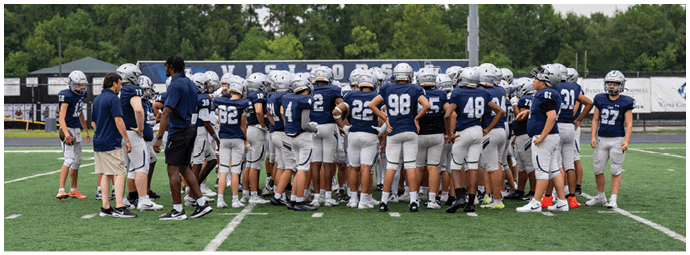 Four people wearing football helmets and white jerseys with “Bishop Verot" written on them stand side by side on a sports field with empty bleachers in the background. Descripci n generada con IA