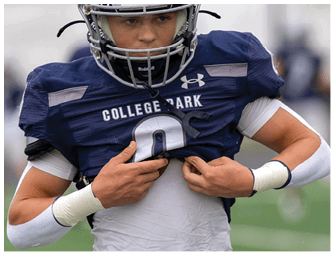 A person wearing a black football helmet and a white and black football jersey is smiling on a sports field with empty bleachers in the background. Descripci n generada con IA