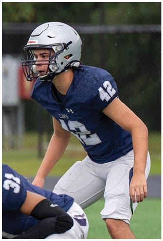 A person wearing a black helmet and a white football uniform with “Bishop Verot" on the jersey is running on a field with another player blurred in the background. Descripci n generada con IA
