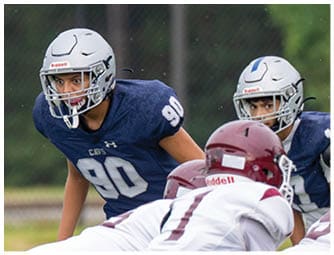 An adult wearing a backward cap adjusts the helmet of a person in a football uniform with the number seven on it during a practice or game. Descripci n generada con IA