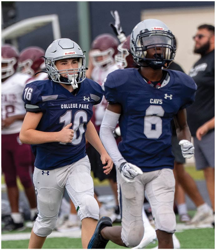 Three people wearing white football uniforms with helmets walk together on a green field during a game or practice. Descripci n generada con IA