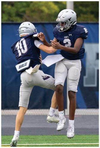 A person wearing a black football helmet and white Bishop Verot jersey with the number 66 stands on a football field with blurred players and bleachers in the background. Descripci n generada con IA