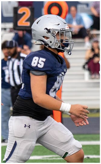 A person wearing a football uniform is kicking a ball on a grassy field with one leg raised high and the other planted on the ground. Descripci n generada con IA