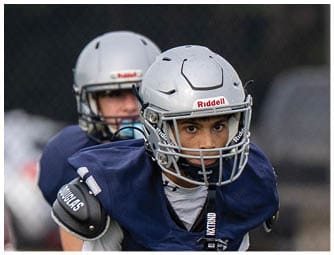 Two people in football uniforms are engaged in a tackle on a field while another player in uniform runs nearby with spectators in the background. Descripci n generada con IA