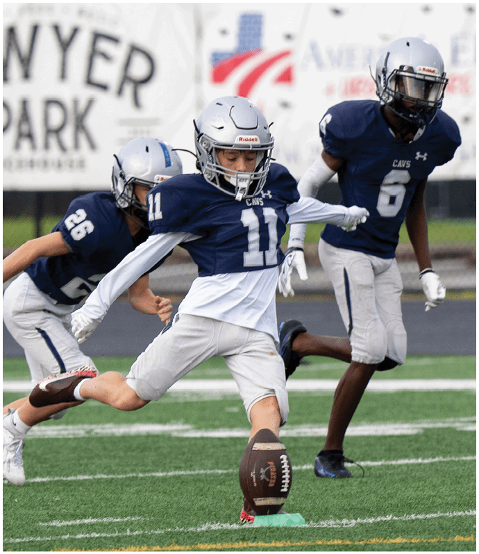 Two people in football uniforms are actively playing on a field at night, with one kicking the ball and the other preparing to block. Descripci n generada con IA