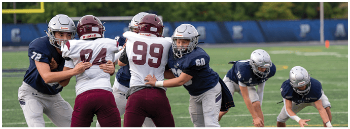 People in football uniforms are actively engaged in a play on a field with bleachers and spectators in the background. Descripci n generada con IA