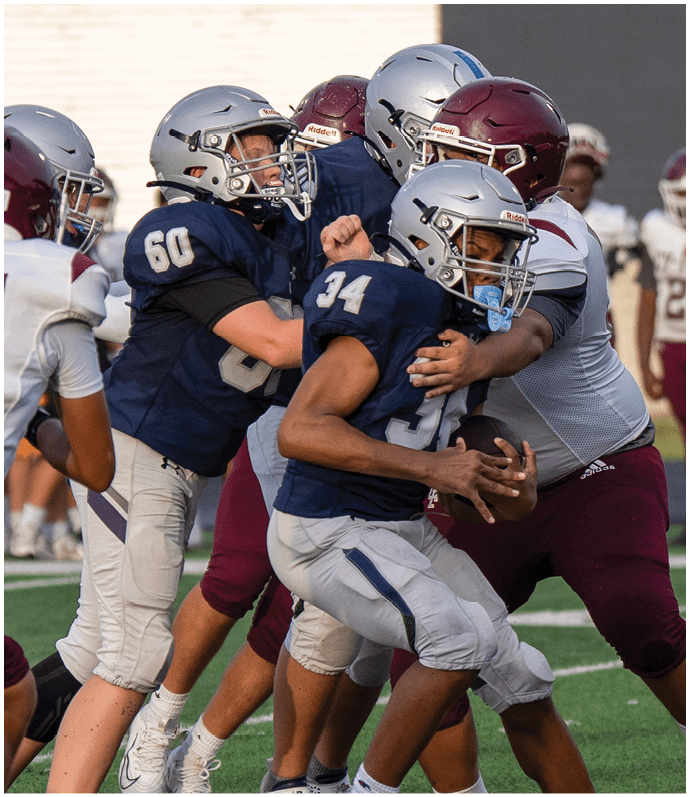 Two people in white sports uniforms with black helmets are running on a football field while two others in green uniforms are nearby. Descripci n generada con IA