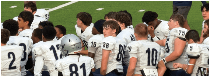 Five people wearing football helmets and uniforms stand on a field facing away from the camera with a group of people in the background. Descripci n generada con IA