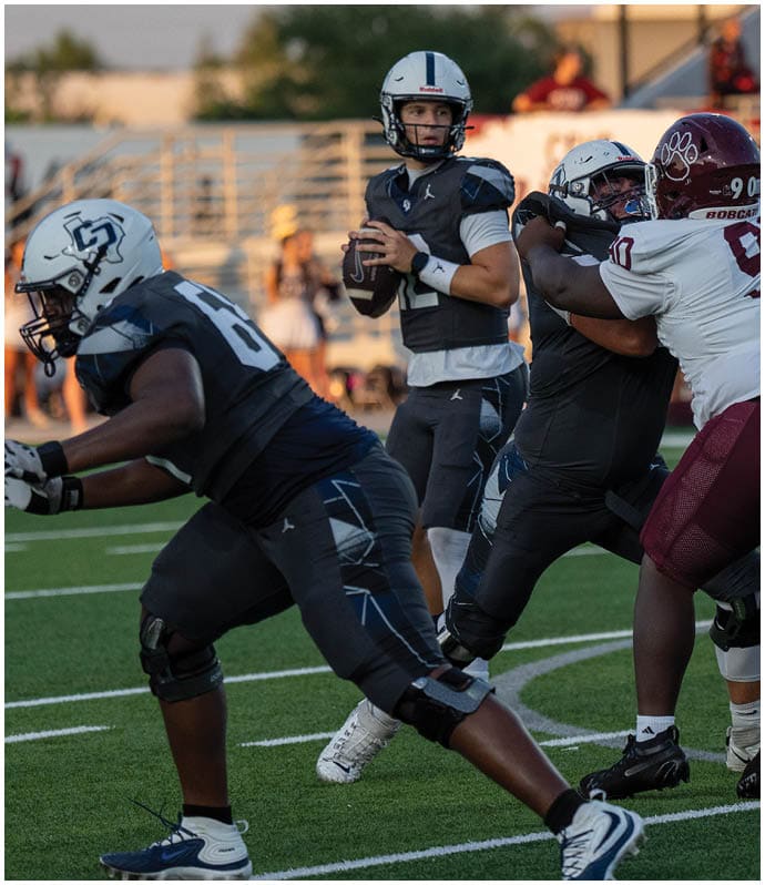 A group of people in football uniforms are engaged in a game on a field with one person holding a football and preparing to throw it. Descripci n generada con IA
