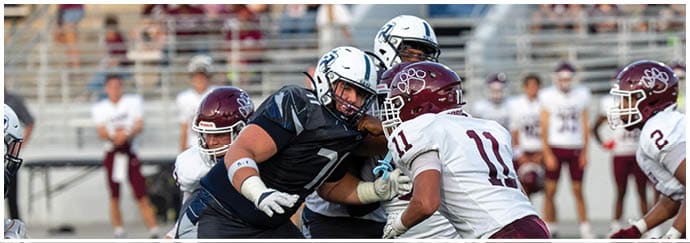 People wearing football helmets and uniforms are engaged in a close physical play on a football field. Descripci n generada con IA