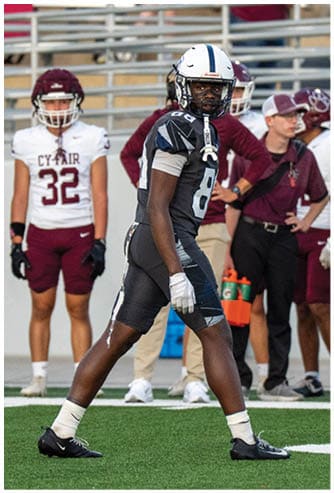 A person in a dark football uniform and helmet is walking on a field with two people in white and maroon football uniforms and others standing in the background. Descripci n generada con IA